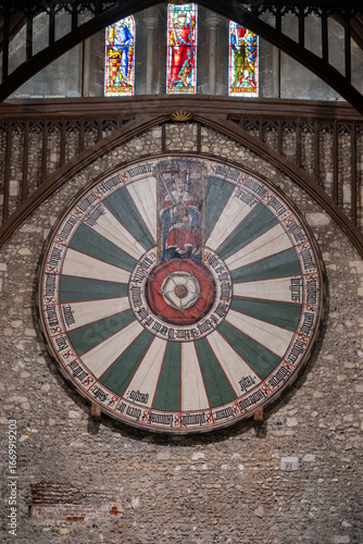 View of the legendary Round Table hanging majestically against the textured stone wall, illuminated by the vibrant stained-glass windows above, Winchester, England, United Kingdom.