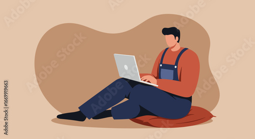Young man in overalls sitting comfortably on a cushion, focused on his laptop, depicting remote work, casual computing, or online learning.