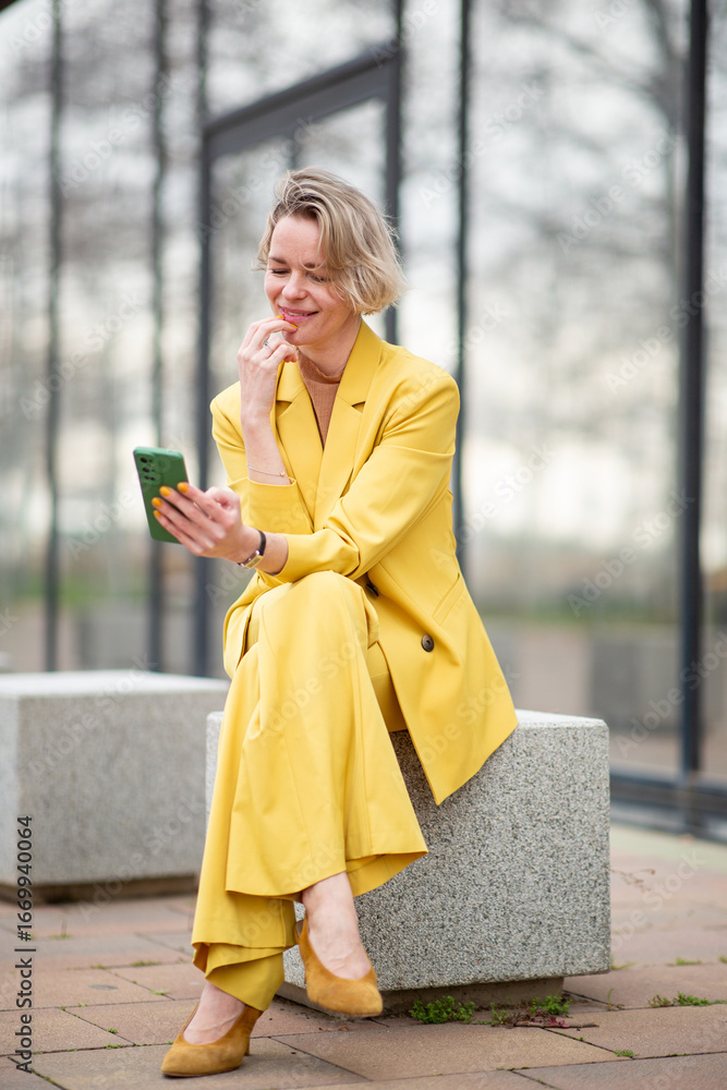 Fototapeta premium Blonde woman in a bright yellow pantsuit sitting on a stone bench, smiling while looking at her mobile phone, enjoying a casual moment outside