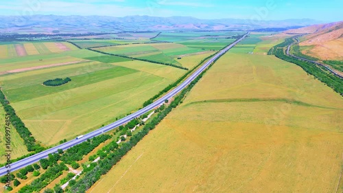 Straight highway cuts through green fields and hills. Aerial view highlights the vastness and order of the rural landscape