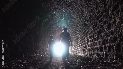 Mother and son walk hand in hand through a dark stone tunnel toward the light. The dramatic lighting and symbolism of moving toward the light create an emotional and inspiring scene