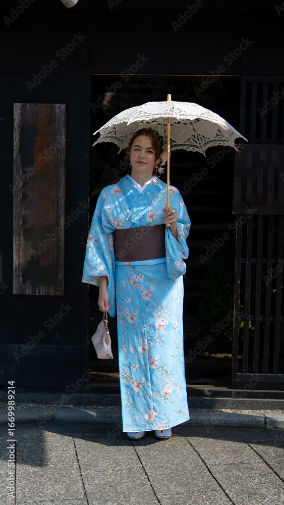 Fototapeta premium A woman in a light blue floral kimono stands at the entrance of a traditional Japanese building, holding a lace parasol and a handbag, blending elegance, heritage, and timeless cultural beauty.