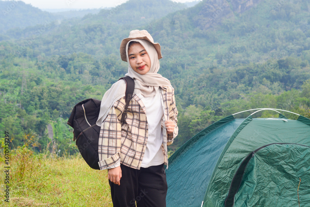 Naklejka premium Sporty Muslim Asian woman standing near tent and backpack, enjoying sunny afternoon in the mountains. Camping lifestyle concept
