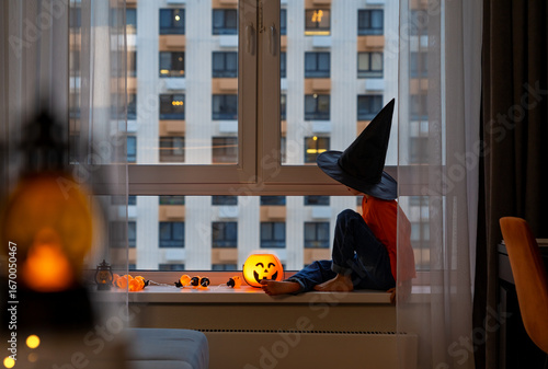 Little girl sits by window in witchs hat and halloween pumpkin lantern in autumn evening. Child at window of big city waits for holiday. Halloween