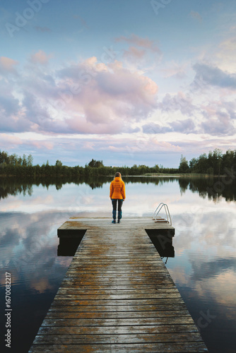 Woman standing alone on pier enjoying sunset lake and forest water reflection view travel outdoor autumn vacations in Finland calm silence scandinavian nature eco tourism
