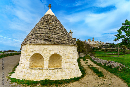 Iconic trullo with Locorotondo skyline in background, Valle d’Itria, Apulia. Traditional conical stone roof house set in the countryside with church domes visible.