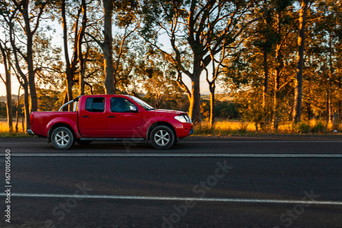 Panning image of red car ute driving past on sealed rural country road in sunset light
