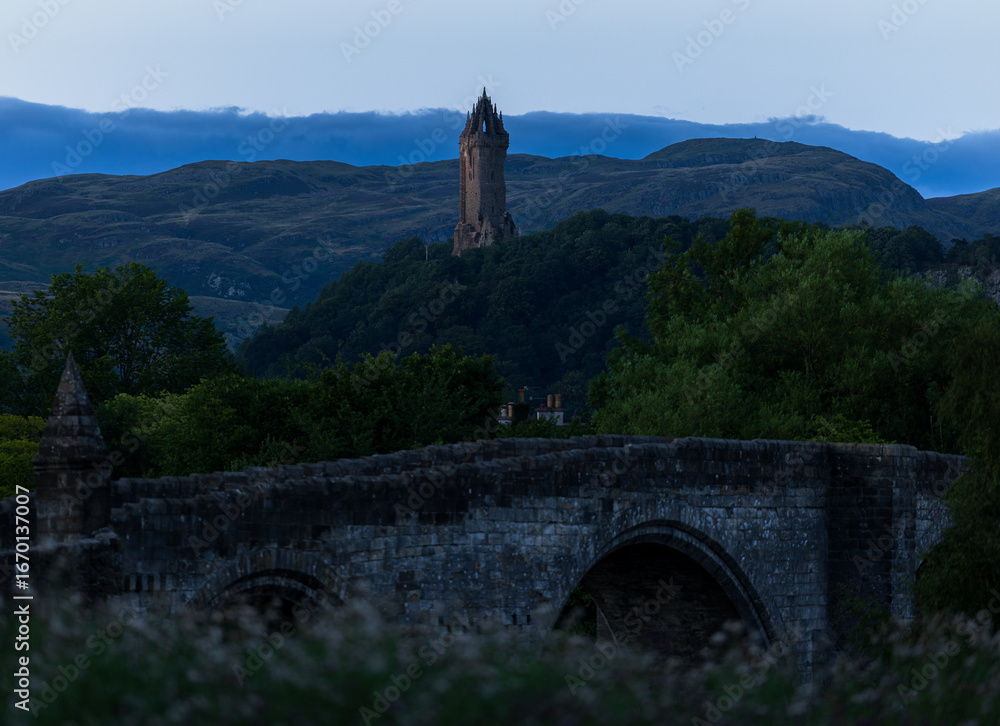 Fototapeta premium Stirling Monument zur blauen Stunde, sanfte Dämmerungsstimmung mit historischer Steinbrücke im Vordergrund und friedlicher Abendatmosphäre in Zentralschottland