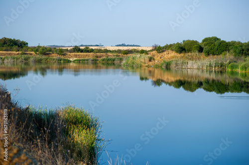 Still waters mirror the soft greenery and golden fields beneath a wide blue sky, evoking rural calm and natural harmony.