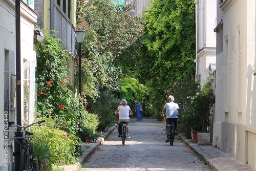 Cyclotourisme dans une ruelle végétalisée en ville, deux cyclistes séniors faisant du vélo en couple dans la verdoyante rue des Thermopyles à Paris, couverte de végétation (France)