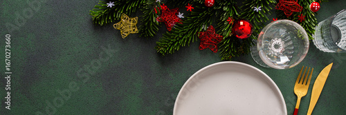 A cozy Christmas table with an empty plate and cutlery on a rich green background. Red Christmas tree decorations and spruse branches complete the festive look, banner