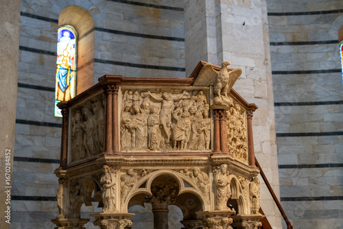 Intricately carved stone pulpit inside a cathedral, supported by ornate columns with sculpted figures and detailed biblical reliefs. A stained glass window and eagle sculpture enhance the rich religio