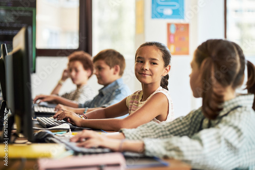 Group of preteen Caucasian and multiethnic children sitting at desks using desktop computers in classroom, girl in foreground turning and smiling at classmate while typing on keyboard