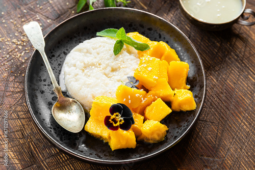 Thai mango sticky rice with coconut milk, garnished with edible flowers on wooden background close up