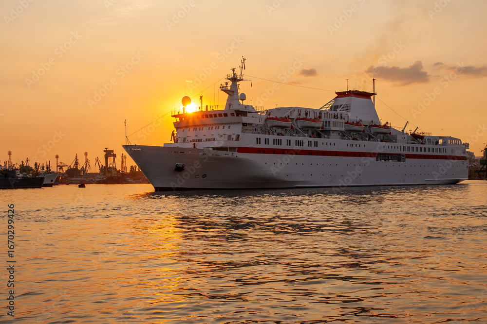 Fototapeta premium Cruise ship sails through the harbor at sunset, reflecting golden hues on the water.