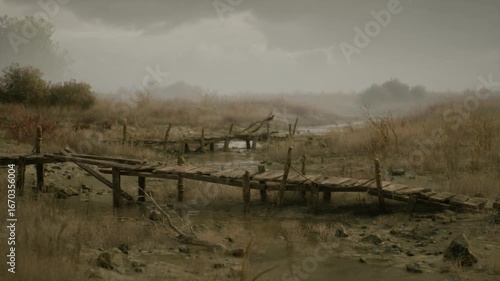 A weathered wooden bridge stretches over muddy terrain, surrounded by dry grasses and sparse trees. The atmosphere is overcast, evoking a somber mood in the landscape.
