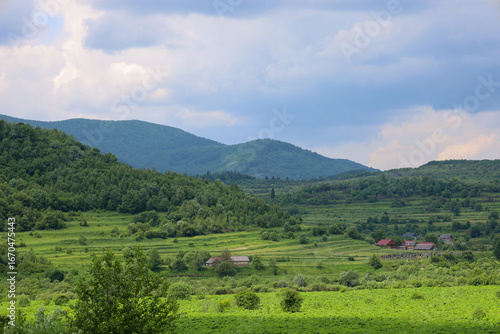 Vast green hills covered with fields and trees stretch towards the horizon. In the valley, scattered rural houses are visible, with gray clouds floating above them, creating an atmosphere of peace and