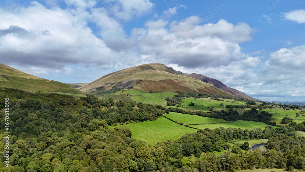 Obraz premium Wide rural landscape of patchwork fields and woodland below a grassy fell under broken cloud in the Lake District, England.