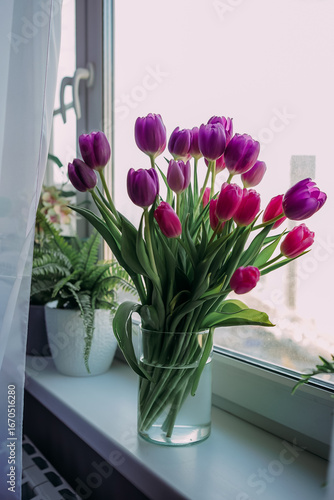 Glass vase with pink and purple tulips on a bright windowsill beside potted plants. Fresh spring flowers and daylight create a cheerful mood, perfect for home decor, floral inspiration