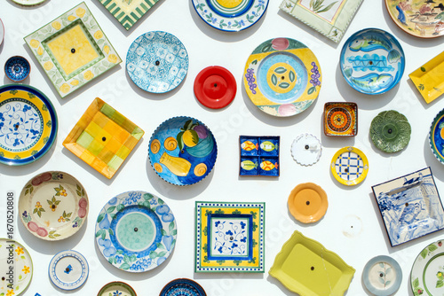 Collection of colorful ceramic plates and dishes with various shapes, patterns, and designs are hanging on a white wall, creating a vibrant and decorative display