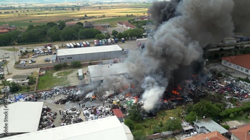 Environmental disaster scene of recycling plant fire. Aerial drone shot shows thick smoke, toxic fumes, burning garbage and firefighters fighting pollution crisis.

