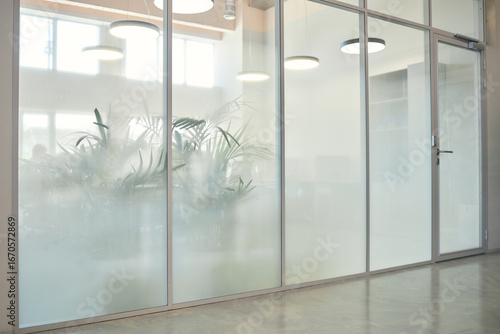 Modern office interior featuring frosted glass wall with large green plants visible behind glass, illuminated by round ceiling lights, empty workspace with closed door