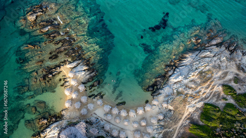 Tranquil turquoise waters gently caress the rocky coastline and sandy beach of Toroni, Greece, dotted with beach umbrellas, creating a serene paradise