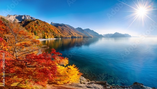a serene autumn day by the sea with golden and red leaves on the mountainsides the calm water reflecting the vibrant colors and the sun shining brightly