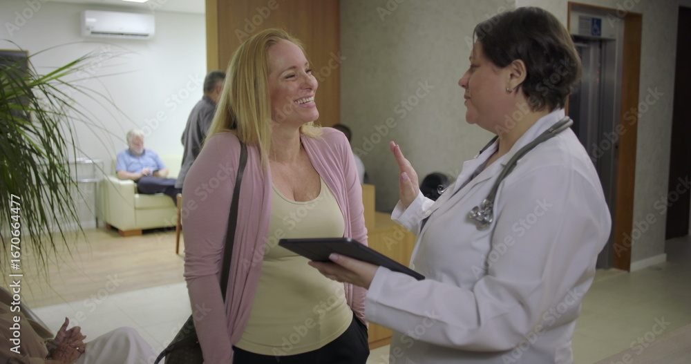 Obraz premium Female doctor reviewing medical details on tablet with woman in reception area of clinic, positive healthcare conversation near waiting area
