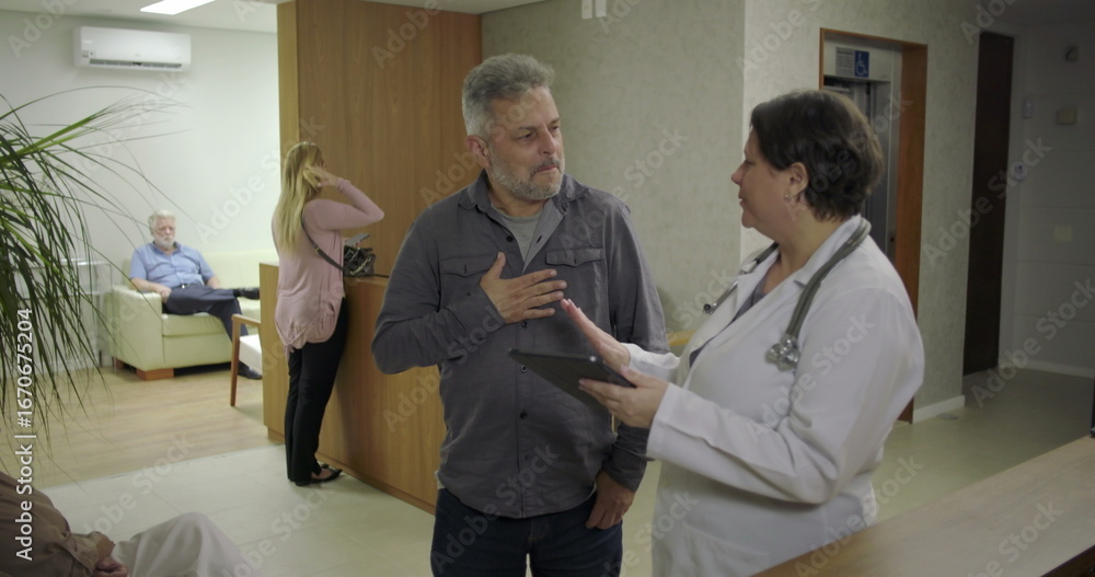 Obraz premium Man expressing relief while speaking with female doctor in clinic hallway, smiling with hand on chest as he receives reassuring news during healthcare discussion