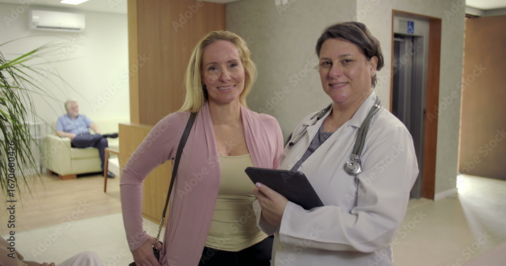 Fototapeta premium Female doctor and patient smiling together in clinic hallway, holding digital tablet near reception area, warm and professional healthcare interaction