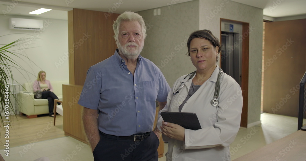 Obraz premium Senior man and female doctor standing together in clinic hallway, facing camera with calm and serious expressions, holding tablet in professional setting