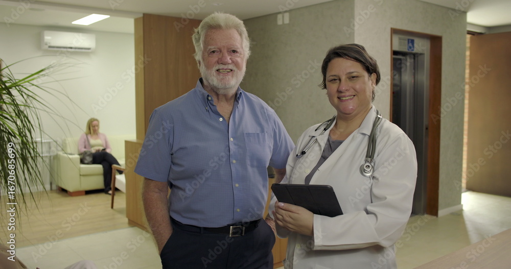 Fototapeta premium Senior male patient and female doctor smiling together in clinic hallway, standing side by side near reception with digital tablet in hand