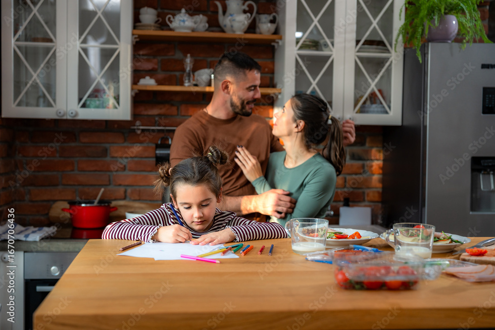 Φωτογραφία While the parents in the background share a tender moment, their daughter focuses intently on her drawing at the table, in the relaxed family setting