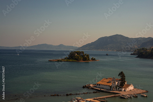 Vlacherna Monastery, located on a small islet near Corfu, Greece, is seen in the foreground with its iconic white buildings and red-tiled roof. The calm blue waters of the Ionian Sea surround it. In t