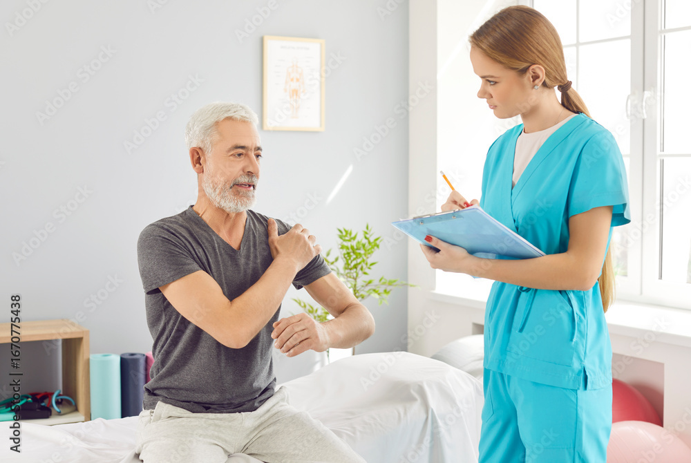 Obraz premium Mature senior man sitting on couch in modern rehab clinic and complaining of pain in the shoulder to a young nurse or doctor physiotherapist. Physiotherapy and physical rehabilitation concept.