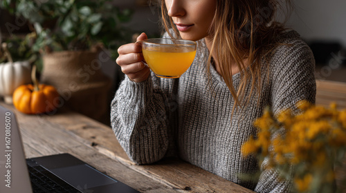 Cozy scene featuring woman enjoying warm drink while working on laptop, perfect for thanksgiving for remote workers. ambiance is inviting with plants and autumn decorations