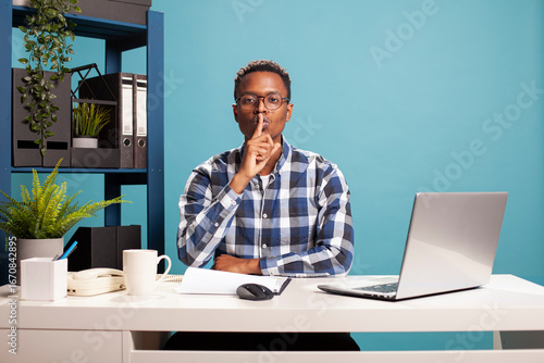 Fotografi Young black man with glasses, sitting at desk and making a hush gesture