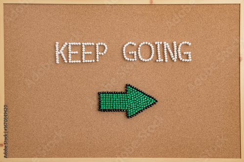 Inspirational message “Keep Going” spelled with push pins on a corkboard, featuring a green arrow pointing right. Symbol of motivation, direction, perseverance, and encouragement.