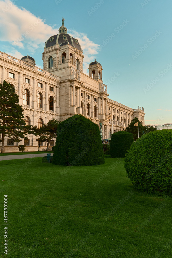 Obraz premium Scenic view of Vienna’s Kunsthistorisches Museum with lush green lawn in the foreground, surrounded by manicured shrubs and pine trees under a blue sky.