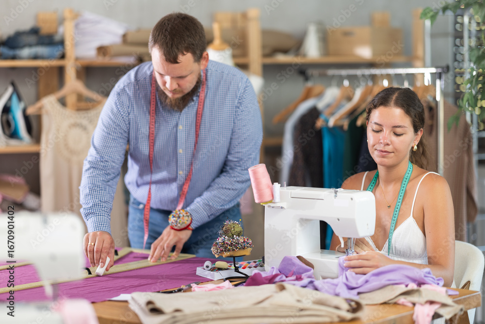 Fototapeta premium Inspired young seamstress working at sewing machine, assembling custom garment from delicate lavender fabric, while male colleague marking fabric with ruler and chalk in modern design studio..