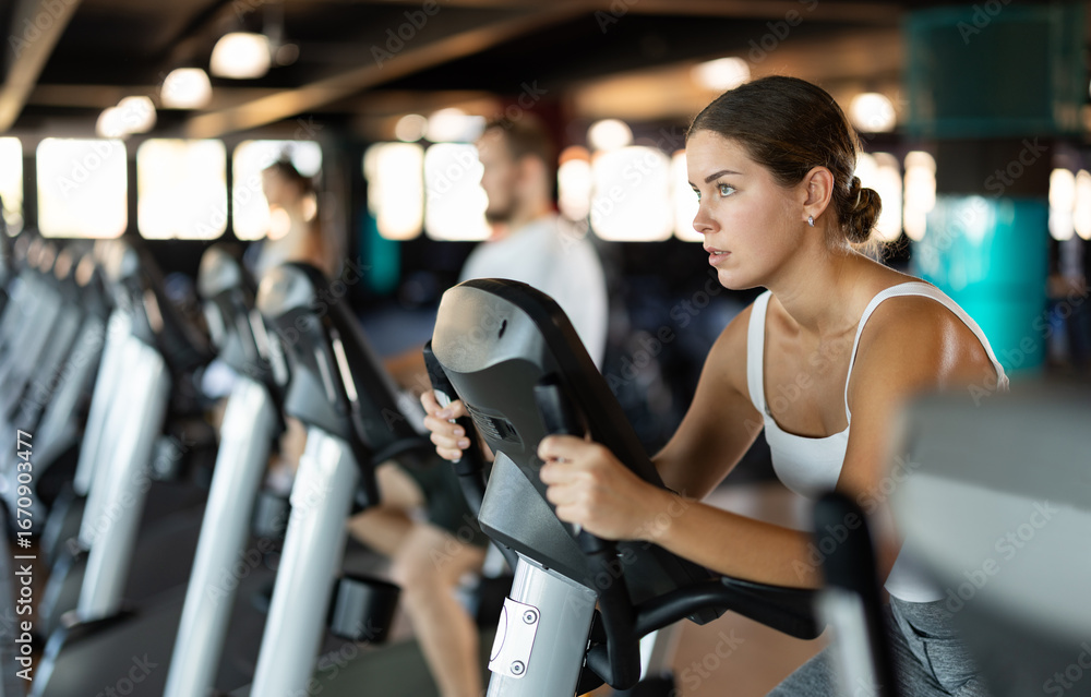 Fototapeta premium Young athletic woman working out on exercise bike in gym