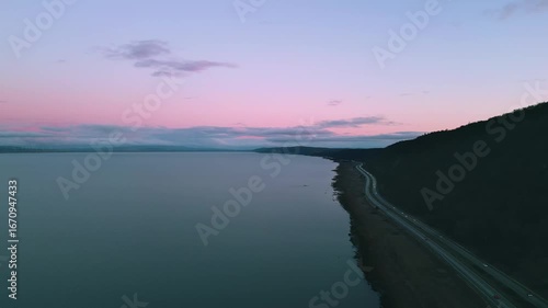 Pink night sky over the lake with highway and cars with headlights on passing below. Wind turbines line the lefthand side of the lake. 