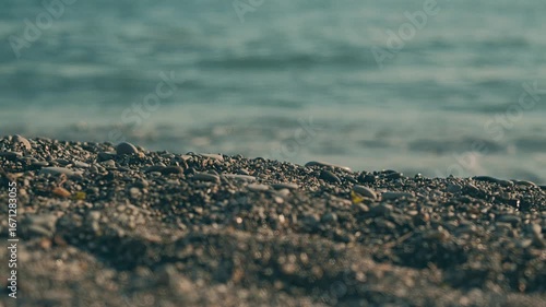 A close-up view of a pebbled beach, with smooth stones dotting the shoreline. The gentle waves of the ocean are blurred in the background, creating a serene atmosphere