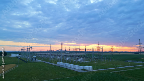Sunset view of a sprawling electrical substation with lush fields and towering pylons, symbolizing sustainable energy infrastructure. Overhead shot highlights technology's intersection with nature.