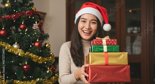Smiling woman in Santa hat holds stack of wrapped Christmas gifts near decorated tree indoors.