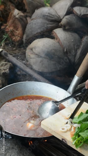 Indonesian woman preparing seblak over charcoal fire in a bamboo saung near rice fields. Traditional cooking method and countryside atmosphere.