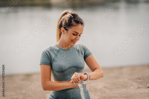 Fotografia Sporty woman checking smartwatch near lake after exercising