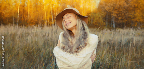Happy smiling mature woman hugs herself in autumn park enjoying warm weather on sunny day outdoors