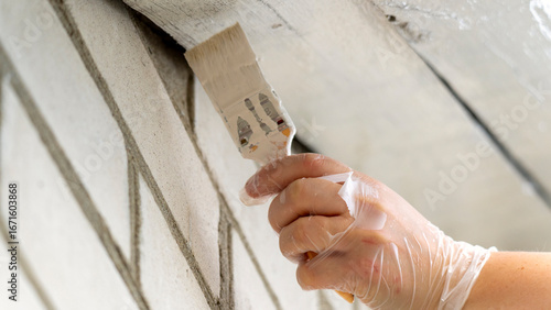 A person is painting a wall with a brush. The wall is made of bricks and the person is wearing gloves